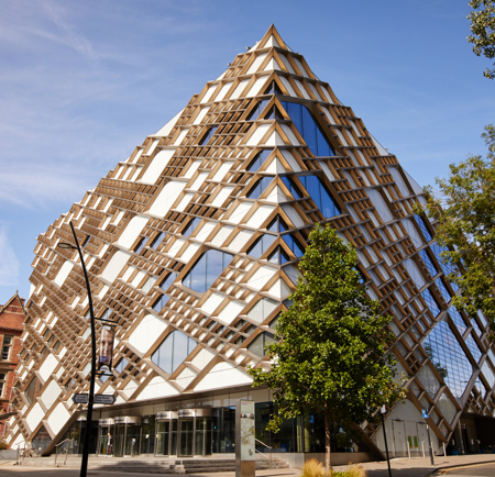 A modern, pyramid-shaped building with a striking geometric design featuring a mix of glass panels and wooden lattice structures. The facade reflects the blue sky, and the building is surrounded by trees and a few older brick buildings on the left. The entrance area has large glass doors, and the street in front includes signage and a lamppost.