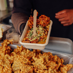 A tray of street food topped with sauce and herbs being held above a counter filled with fried items.