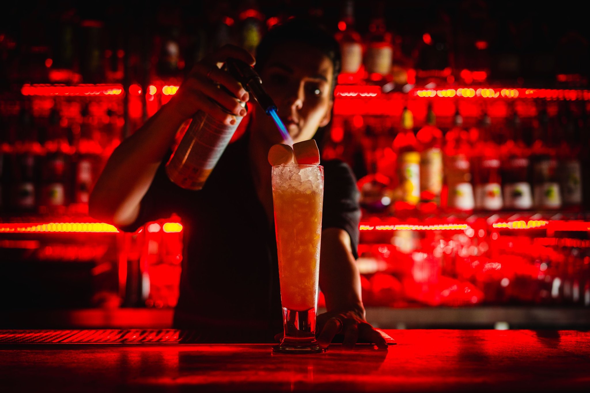 A man mixing a cocktail in a bar that has subdued red lighting.