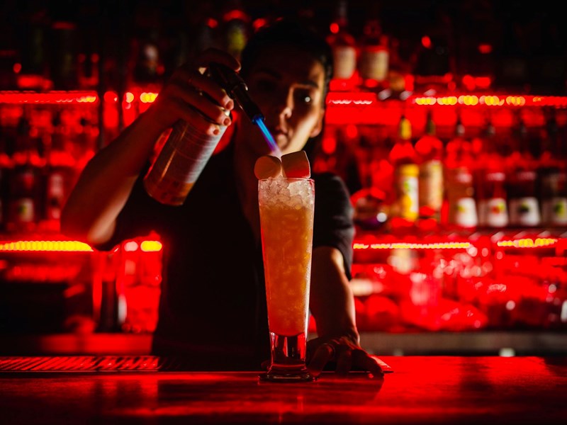 A man mixing a cocktail in a bar that has subdued red lighting.