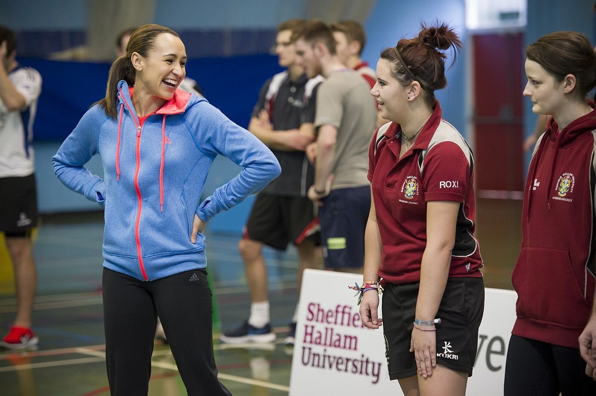 A group of people standing indoors on a sports court, wearing athletic clothing. One person in the foreground is dressed in a bright blue zip-up hoodie with pink accents and black leggings, while others wear maroon sports shirts with logos and black shorts. A white sign with red text reads “Sheffield Hallam University.” The background shows more individuals in casual sportswear and gym flooring with court markings.