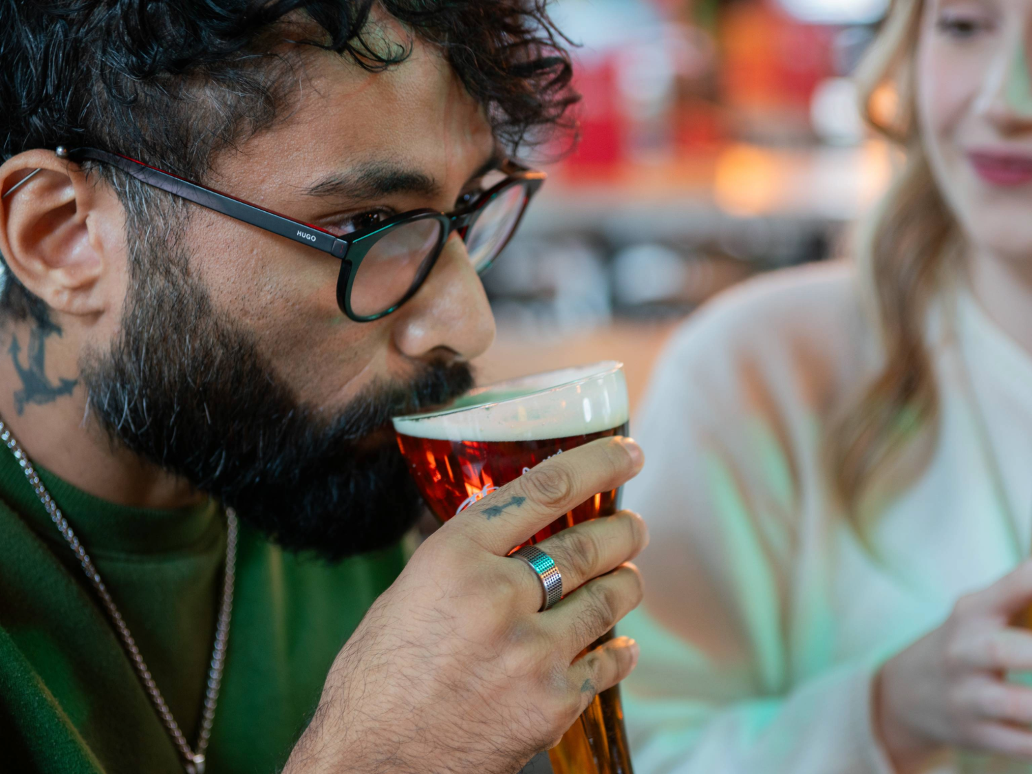 Close-up of a person holding a pint of beer with a thick foamy head in a clear glass. The individual is wearing a green top and a silver necklace, with a silver ring visible on their hand. Another person in a white top is partially visible beside them. The background shows a bar area with warm lighting and blurred bottles, creating a casual indoor setting.