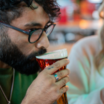 Close-up of a person holding a pint of beer with a thick foamy head in a clear glass. The individual is wearing a green top and a silver necklace, with a silver ring visible on their hand. Another person in a white top is partially visible beside them. The background shows a bar area with warm lighting and blurred bottles, creating a casual indoor setting.