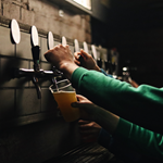 A row of beer taps on a wall. Several people are pouring pints of beer.