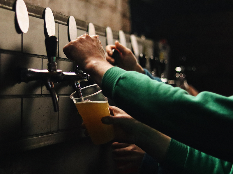 A row of beer taps on a wall. Several people are pouring pints of beer.