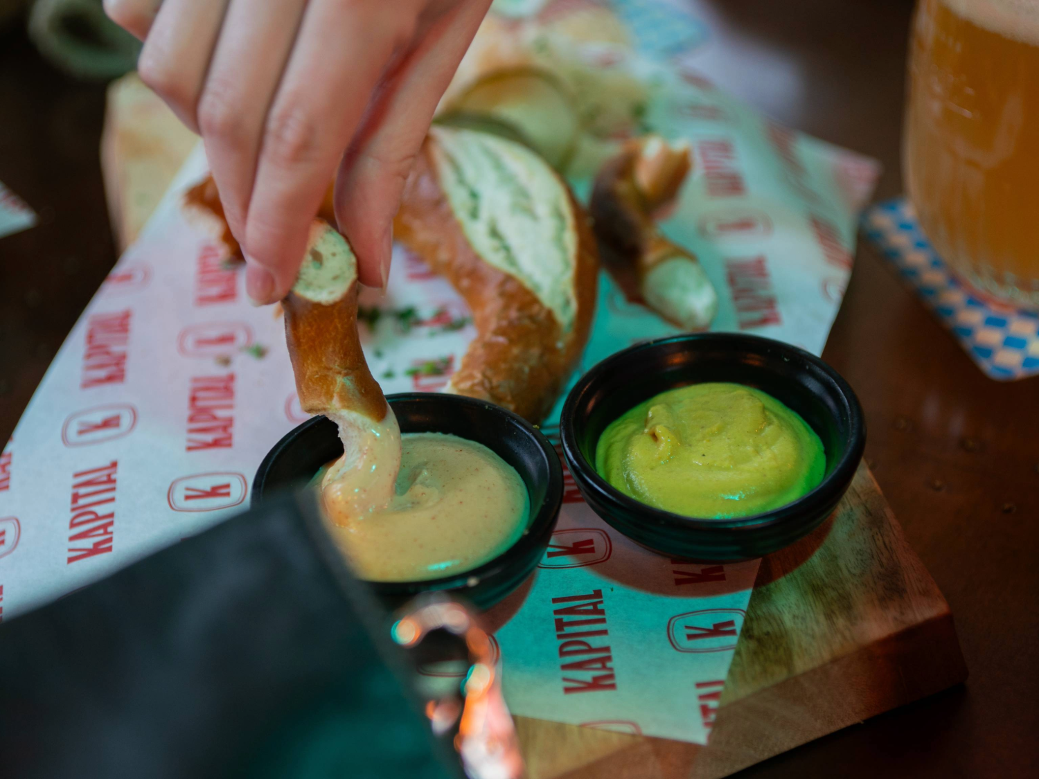 A hand dips a piece of pretzel into a creamy mustard sauce in a black bowl, with another bowl of yellow mustard beside it. The food is served on a wooden board lined with branded Kapital paper, featuring more pretzel pieces in the background. A glass of beer with a foamy head is partially visible to the right on a blue-and-white checkered coaster, creating a casual bar setting.