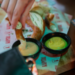 A hand dips a piece of pretzel into a creamy mustard sauce in a black bowl, with another bowl of yellow mustard beside it. The food is served on a wooden board lined with branded Kapital paper, featuring more pretzel pieces in the background. A glass of beer with a foamy head is partially visible to the right on a blue-and-white checkered coaster, creating a casual bar setting.