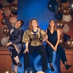 A photo of three women sat on the edge of a stage.
