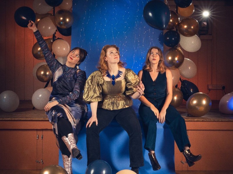 A photo of three women sat on the edge of a stage.