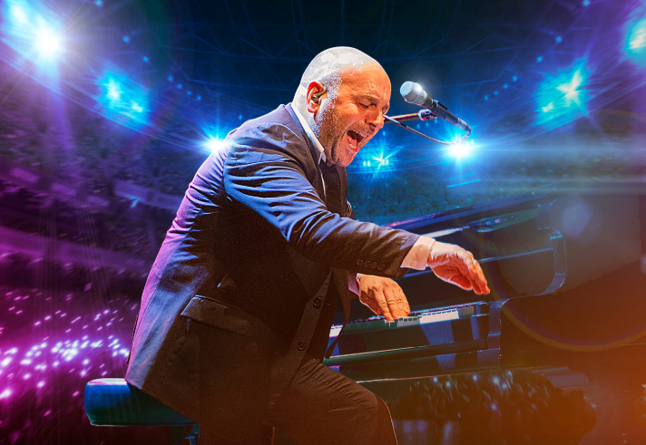 A performer in a dark suit is seated at a grand piano on a brightly lit stage. The musician is leaning forward with one arm extended toward the keys, while a microphone is positioned above the piano. The background shows vivid blue and purple stage lights and a large audience area illuminated with glowing lights.