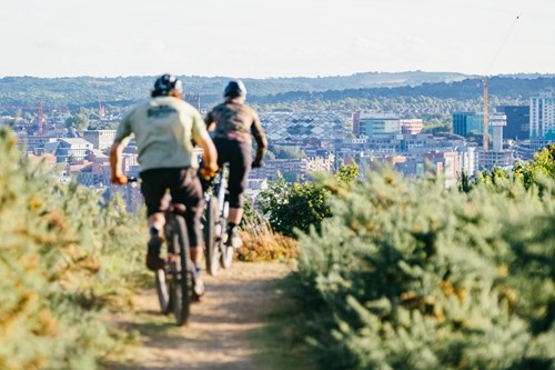 Two cyclists riding mountain bikes along a dirt trail surrounded by green shrubs, with a panoramic view of a city skyline and hills in the background under a clear sky.