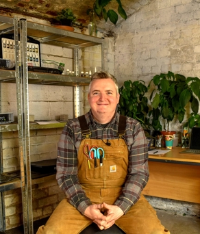 A man, in overalls, sat in a workshop smiling at the camera.