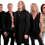 Five people standing together in a studio setting, dressed in rock-style outfits. Clothing includes black leather pants, open shirts, long coats, and patterned vests. Several wear necklaces with cross pendants. The background is plain white, and the group is posed in a staggered formation.