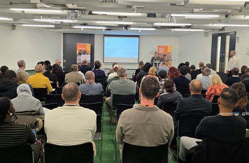 A large group of people seated in a conference room listening to a speaker at a podium. The presentation slide on the screen reads “How to fund a business: grants, loans and investment,” and two orange Business Sheffield banners stand on either side of the room.