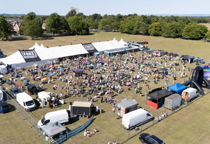 A large outdoor music festival set in an open grassy field with a white marquee tent labeled “FAKE FESTIVALS.” Crowds of people are gathered in front of the tent, sitting on blankets or standing near the stage area. Surrounding the festival are food stalls, trucks, and inflatable attractions, with trees and countryside visible in the background under a clear blue sky.