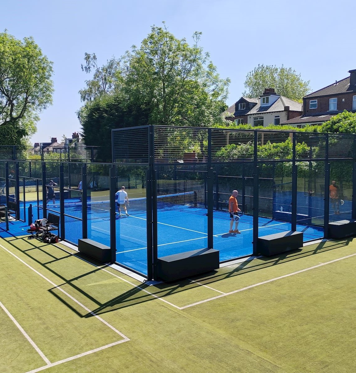 Outdoor padel courts with bright blue playing surfaces enclosed by glass and metal fencing, set within a larger green tennis court area. Several players are on the padel courts, holding rackets and playing under clear blue skies. Surrounding the courts are tall trees and residential houses, creating a leafy suburban backdrop. Sports bags and equipment are placed near the court entrance.