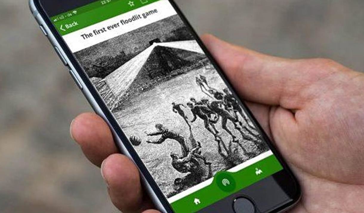 A hand holding a smartphone with the Home of Football: Walking Tour App on the screen.