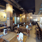 The interior of Cubana Tapas Bar. Interior of a rustic-style restaurant featuring wooden tables and chairs arranged in rows, each set with menus and condiments. A long wooden bar with high stools runs along the right side, with a counter displaying various items. The walls have a textured, warm-toned finish with framed pictures and decorative elements. Overhead, dark exposed beams and recessed lighting create a cozy atmosphere.