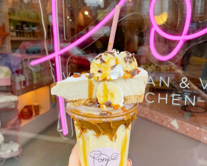 Hand holding a dessert drink topped with a slice of pie, banana slices, whipped cream, and caramel drizzle. The drink is in a clear cup with a pink straw and a label reading ‘Pom.’ In the background, a shop window with pink neon signage and shelves of colorful items is visible.