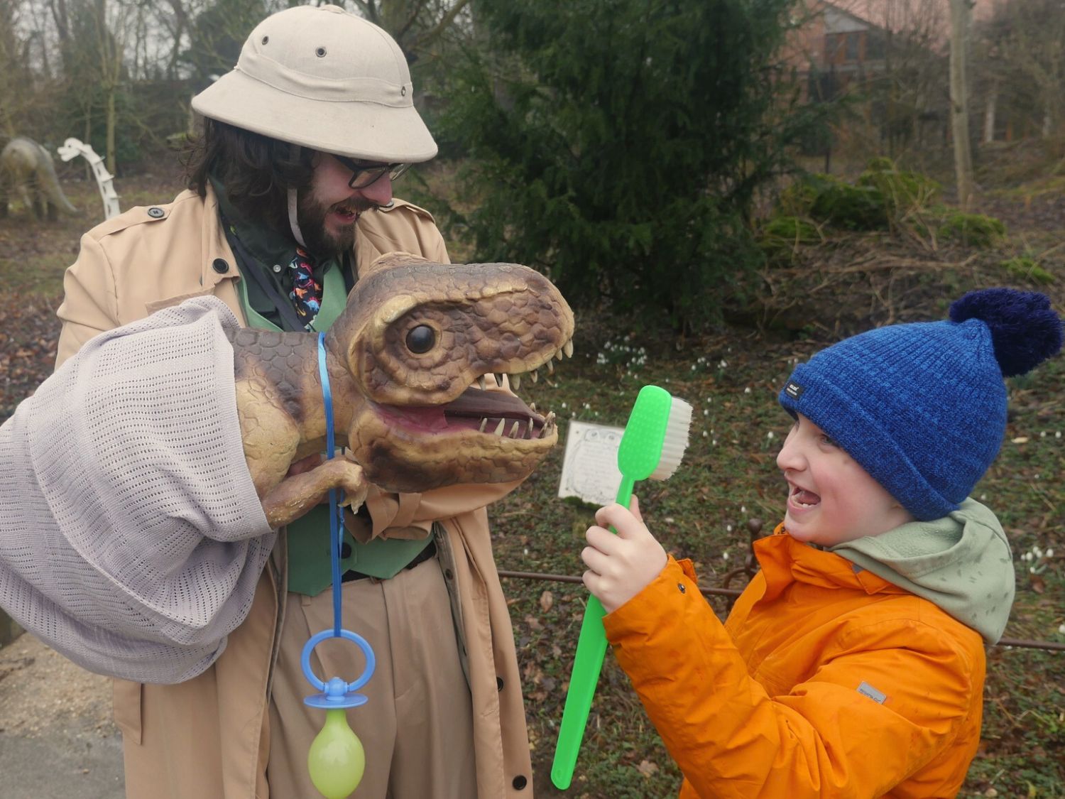 A child looking at a puppet dinosaur, being operated by a man in a pith helmet.