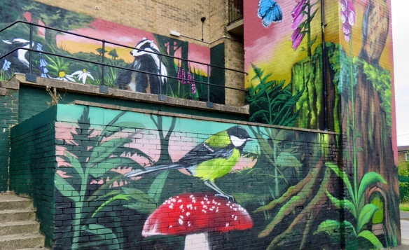 Colorful nature-themed mural painted on the side of a building, featuring a green and yellow bird perched on a red mushroom, surrounded by lush green plants. Additional artwork includes badgers among flowers, a blue butterfly, and tall pink foxgloves against a sunset gradient background.