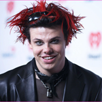 Person wearing a black leather jacket and layered necklaces, including a padlock pendant, with a black scarf tied around the neck. Hair is styled in spiky red and black strands with sunglasses resting on top. The background features a white backdrop with red heart-shaped logos.