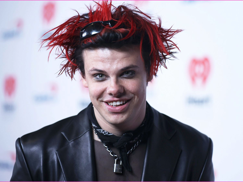 Person wearing a black leather jacket and layered necklaces, including a padlock pendant, with a black scarf tied around the neck. Hair is styled in spiky red and black strands with sunglasses resting on top. The background features a white backdrop with red heart-shaped logos.