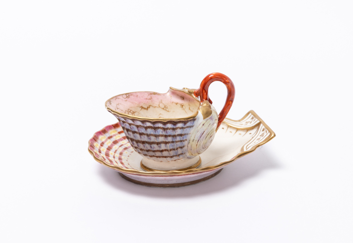 A decorative ceramic cup and saucer designed to resemble seashells are displayed against a plain white background. The cup has a scalloped, shell‑like form with textured ridges, soft pink and cream tones inside, and a curved, coral‑coloured handle. The saucer also mimics a shell shape with patterned ridges and a mix of cream, red, and beige accents, complementing the cup’s intricate, coastal-inspired design.
