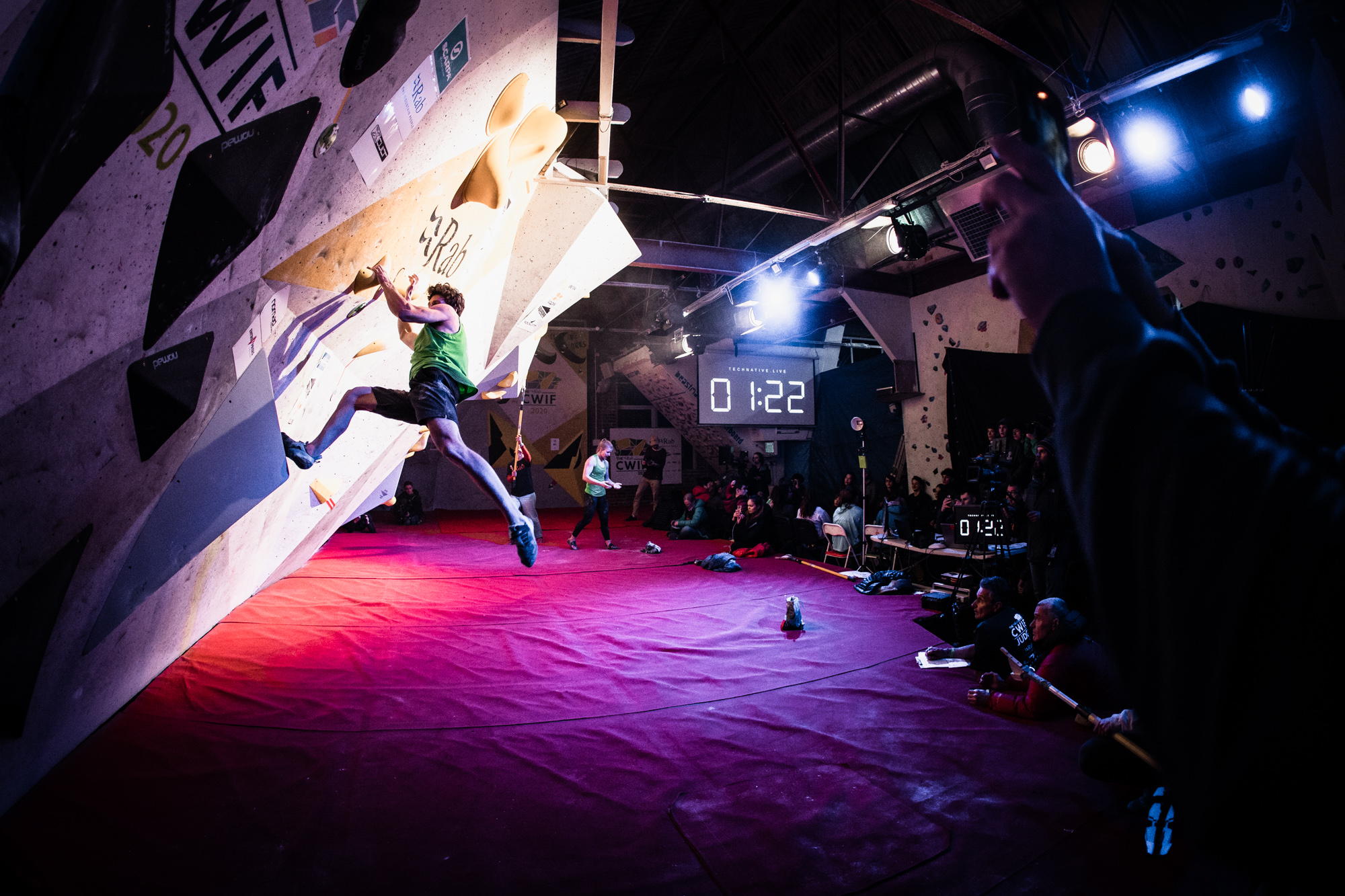 A man is climbing on a wall. He has two hands and one foot on the wall, with one foot mid-climb in the air. There is a digital timer in the background displaying one minute and twenty two seconds.