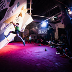 A man is climbing on a wall. He has two hands and one foot on the wall, with one foot mid-climb in the air. There is a digital timer in the background displaying one minute and twenty two seconds.