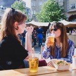 Two women in sunglasses sit drinking a pint of beer with food stall in the background 