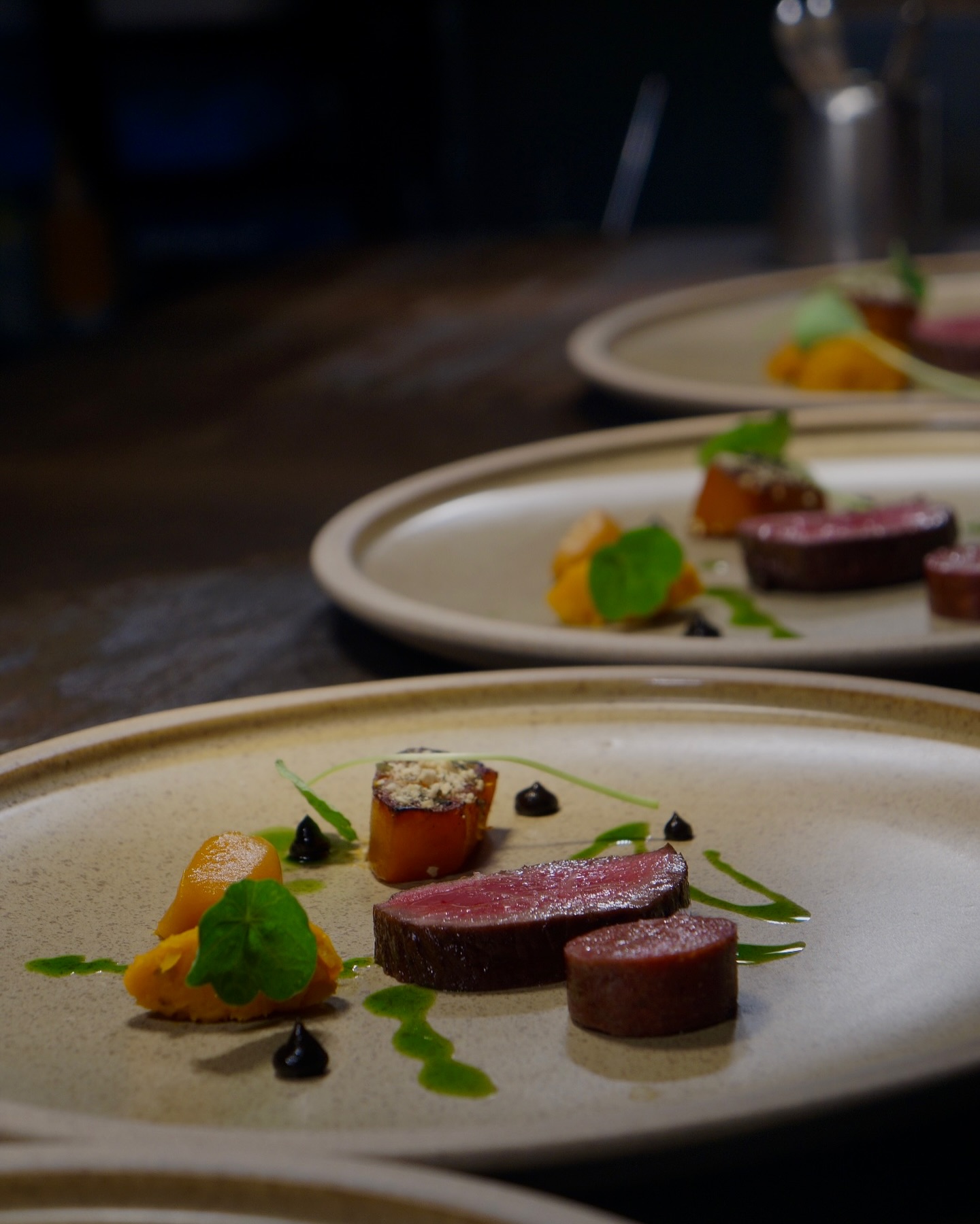 Close-up of a plated fine-dining dish featuring slices of seared meat, cylindrical cuts, bright orange vegetables, green herb leaves, and artistic drizzles of sauce on a neutral-toned plate.