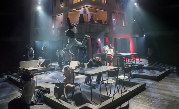 A dramatic stage performance featuring multiple actors in an industrial-style set. The foreground shows scattered tables and chairs with performers in dynamic poses, including one person leaping into the air. The background includes a two-level structure with additional actors visible on the upper floor, illuminated by theatrical lighting in shades of blue and red. The scene conveys high energy and movement.