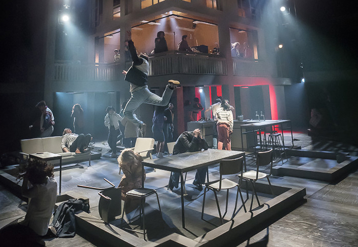 A dramatic stage performance featuring multiple actors in an industrial-style set. The foreground shows scattered tables and chairs with performers in dynamic poses, including one person leaping into the air. The background includes a two-level structure with additional actors visible on the upper floor, illuminated by theatrical lighting in shades of blue and red. The scene conveys high energy and movement.