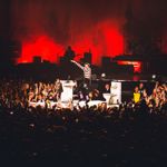 A performer on an elevated stage reaches out toward a tightly packed crowd under red lighting. Musicians and equipment are visible on the stage behind them.