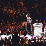 A performer walks along a narrow platform surrounded by fans holding phones and reaching toward the stage. Stage props resembling tiled bathroom fixtures are visible nearby.