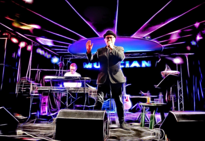 Two people performing on stage, one standing and singing and the other sat playing a keyboard.