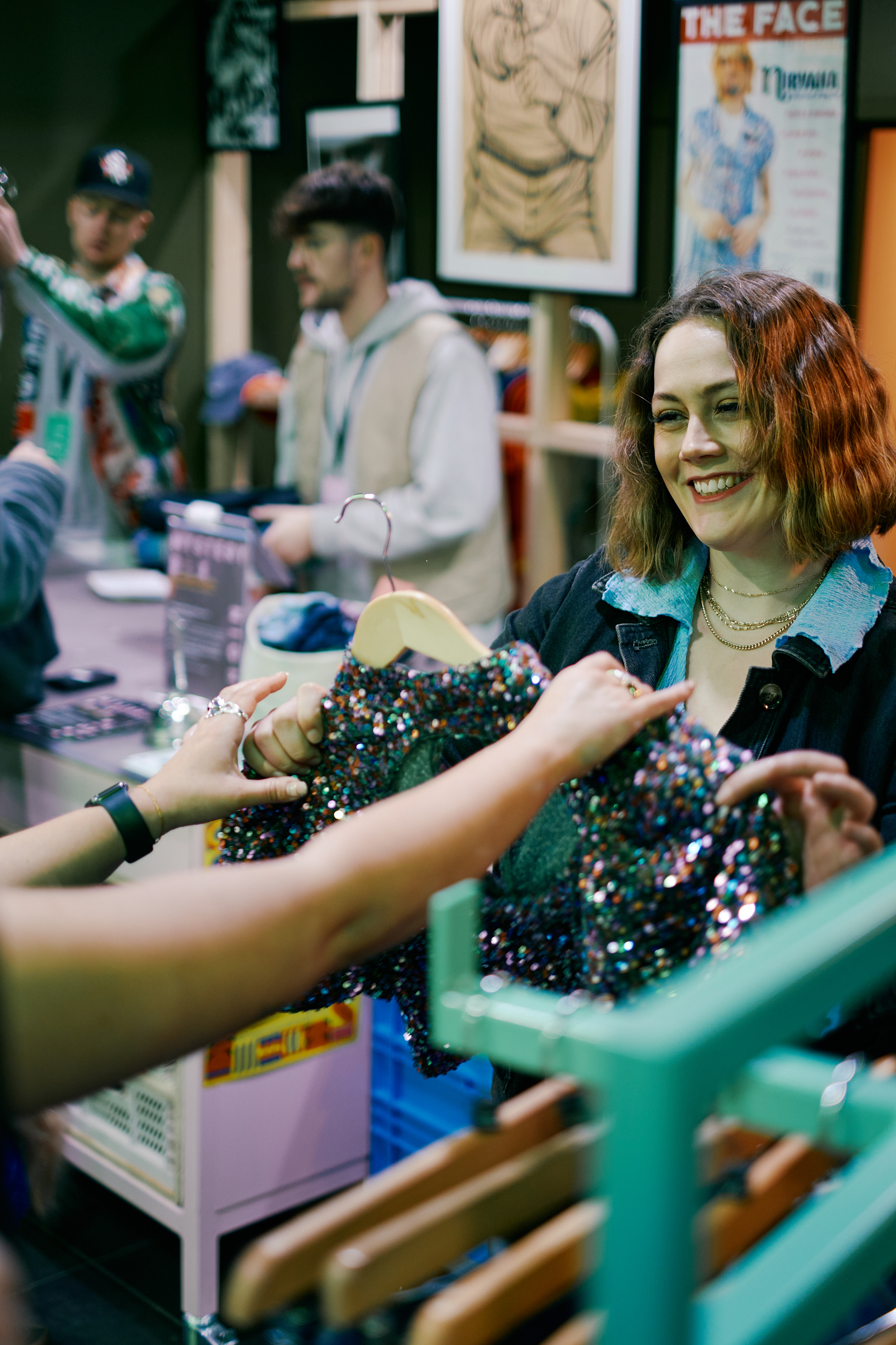 Two women admire a sequined top at a vintage clothes shop. There are other shoppers in the background.