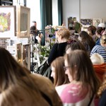 A busy indoor market or fair with people browsing stalls selling artwork, crafts, and plants. A framed painting of a house with colorful foliage is prominently displayed. The scene captures a lively community event focused on local art and handmade goods.