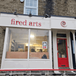 Exterior of Fired Arts ceramics studio with white frontage, red door, and large front window showing the studio interior.