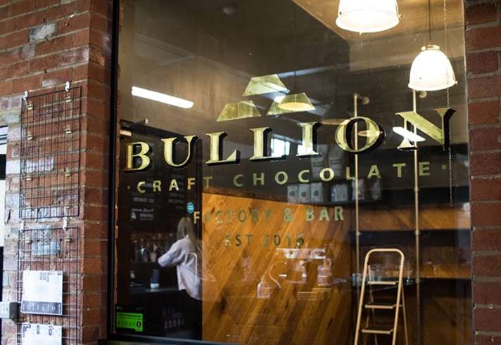 Storefront of Bullion Craft Chocolate, featuring a glass window with the gold Bullion logo and name. Inside, wooden shelves, a ladder, and hanging lights are visible. A person is seen through the window. The building has a red brick exterior.
