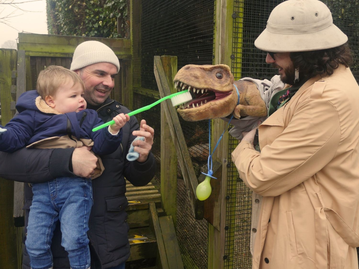 A small child looking at a puppet dinosaur, being operated by a man in a pith helmet.