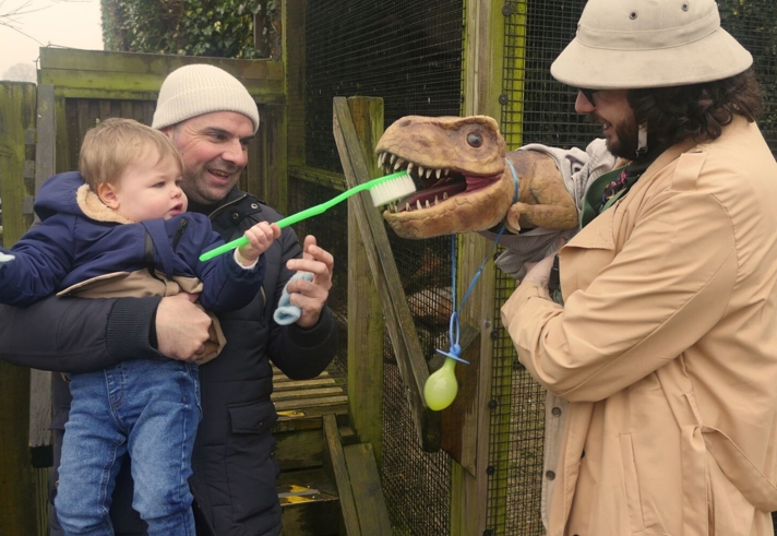A small child looking at a puppet dinosaur, being operated by a man in a pith helmet.
