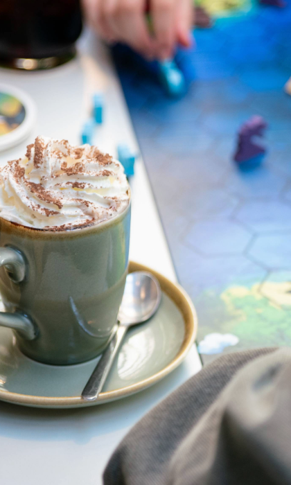 A close-up of a cappuccino sitting on a table, next to a board game. A hand is moving one of the pieces from the game. 