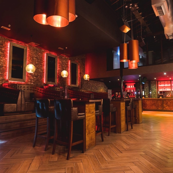 Interior shot of Crystal. Modern bar interior with warm lighting, exposed brick walls, and wooden herringbone flooring. The space features high tables with leather chairs, booth seating along the wall, and a long bar counter with illuminated shelves stocked with bottles. Copper pendant lights hang from the ceiling, adding to the stylish atmosphere.