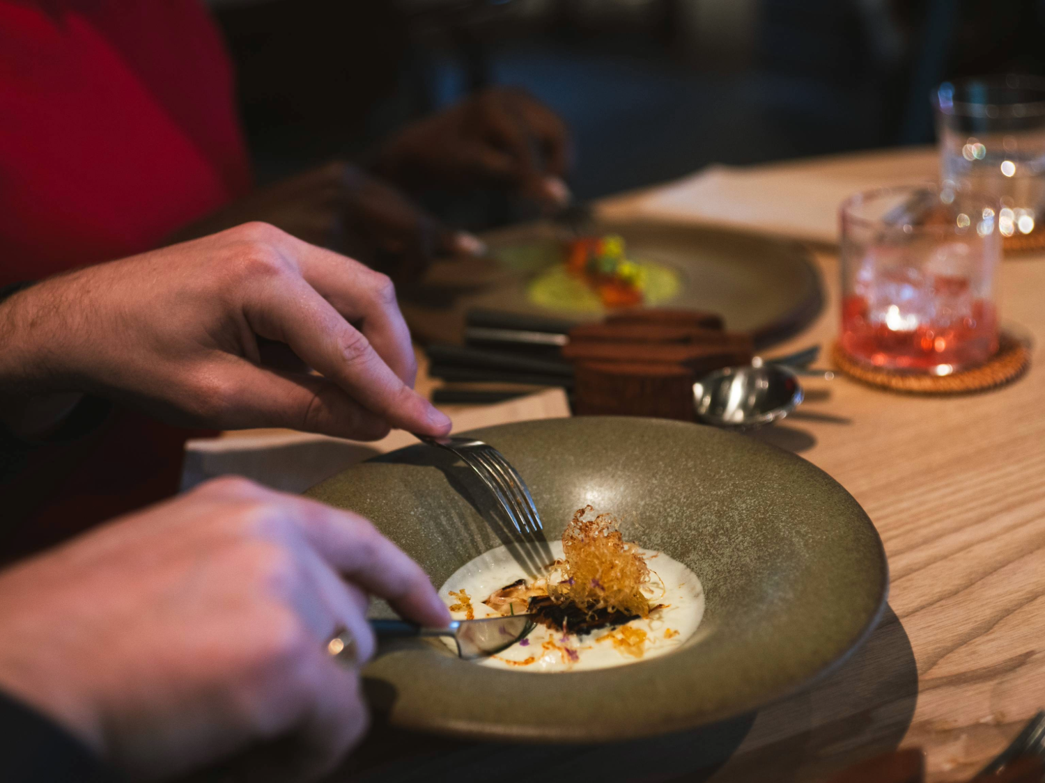 Close-up of two people dining at a wooden table in a modern restaurant. One person is using a fork and knife to eat a dish served on a green ceramic plate, featuring a creamy white sauce and a crispy garnish. Another plate with a colourful dish is visible in the background, along with cutlery and a glass containing a pink drink on a coaster. The setting is warm and elegant, with a focus on fine dining presentation.