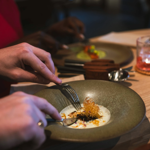 Close-up of two people dining at a wooden table in a modern restaurant. One person is using a fork and knife to eat a dish served on a green ceramic plate, featuring a creamy white sauce and a crispy garnish. Another plate with a colourful dish is visible in the background, along with cutlery and a glass containing a pink drink on a coaster. The setting is warm and elegant, with a focus on fine dining presentation.