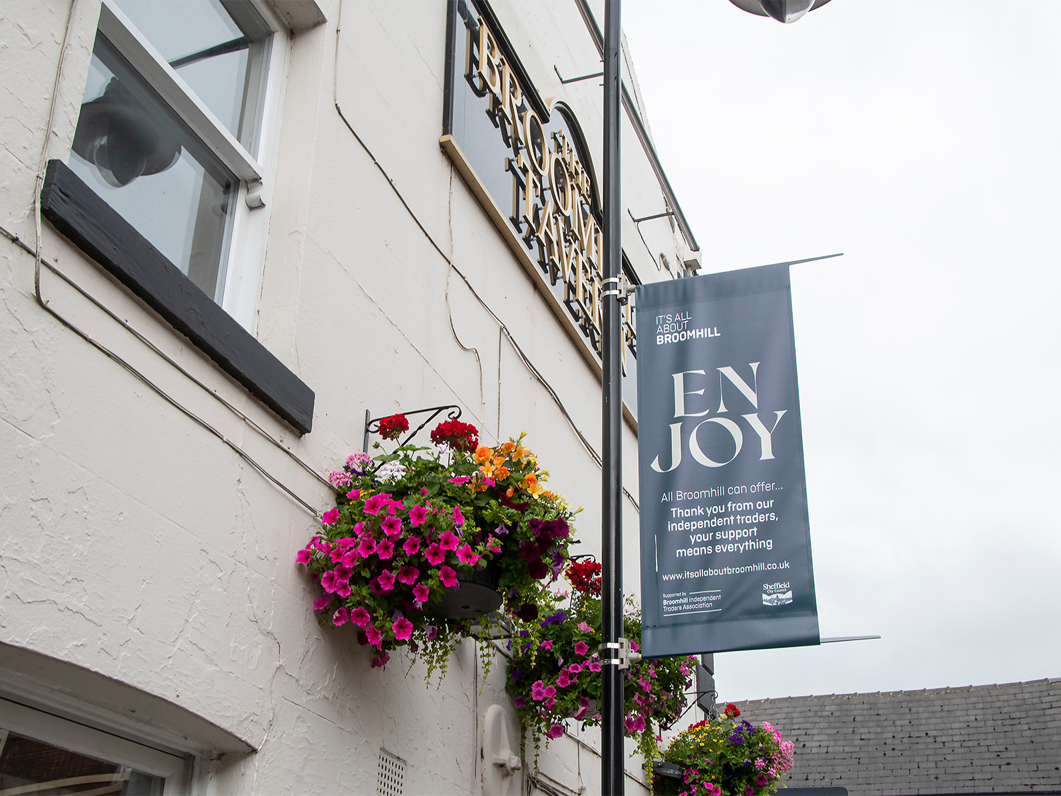 Exterior wall of a light-colored building with hanging baskets filled with vibrant flowers in pink, purple, and red. A vertical banner attached to a pole reads “ENJOY” and promotes local independent traders in Broomhill, with a website link at the bottom. Part of the building sign for “Broomhill Tavern” is visible above the baskets, and rooftops appear in the background under a cloudy sky.