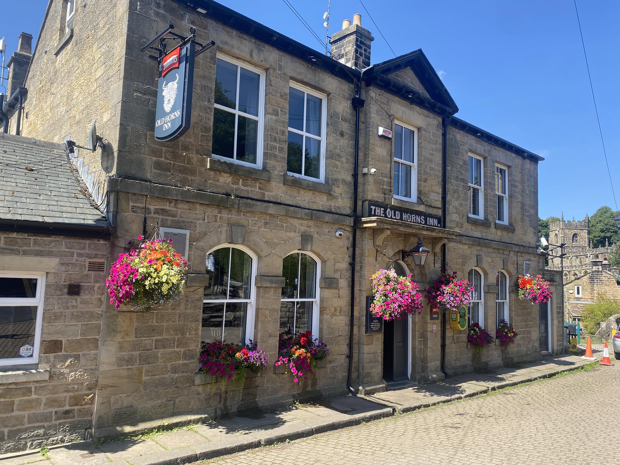The Old Horns Inn photographed on a sunny day, showcasing its traditional stone façade, arched windows, colourful hanging baskets and cobbled street setting.