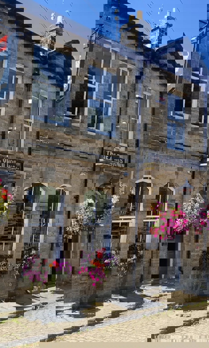 The Old Horns Inn photographed on a sunny day, showcasing its traditional stone façade, arched windows, colourful hanging baskets and cobbled street setting.